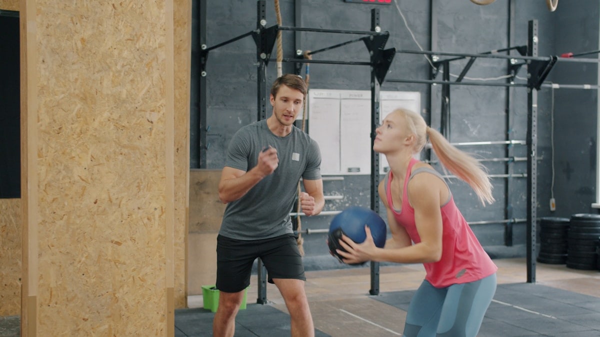 Young woman is exercising with ball in gym while personal coach is motivating girl talking giving advice. Training and active lifestyle concept.